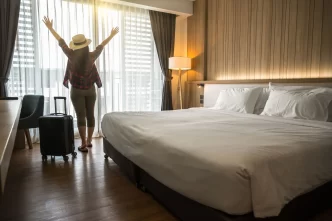 Cliente in piedi vicino al letto in una camera d'albergo, con le braccia alzate, accanto a una valigia.