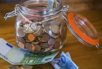 Salvadanaio di vetro pieno di monete con banconote euro sul tavolo.