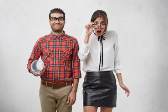 Uomo sorridente in camicia a quadri e donna sorpresa con occhiali, su sfondo chiaro.