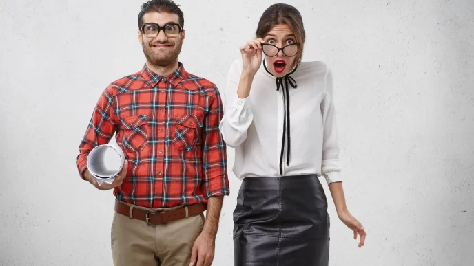 Uomo sorridente in camicia a quadri e donna sorpresa con occhiali, su sfondo chiaro.