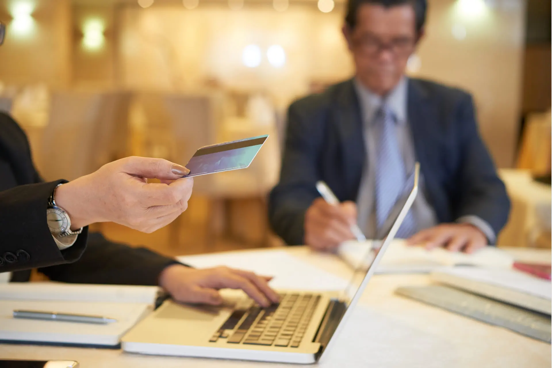 Persona utilizza una carta di credito vicino a un laptop, con un collega che scrive appunti sullo sfondo.