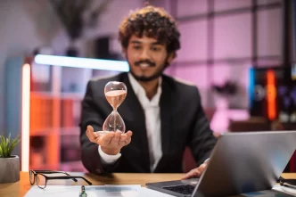 Uomo d'affari sorridente in abito formale seduto alla scrivania con un computer portatile e tenendo la clessidra in mano.