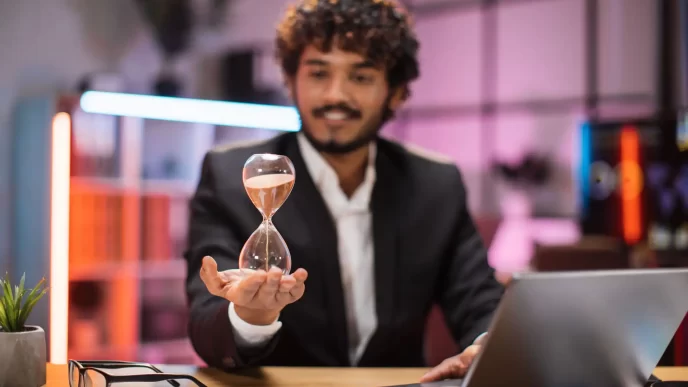 Uomo d'affari sorridente in abito formale seduto alla scrivania con un computer portatile e tenendo la clessidra in mano.