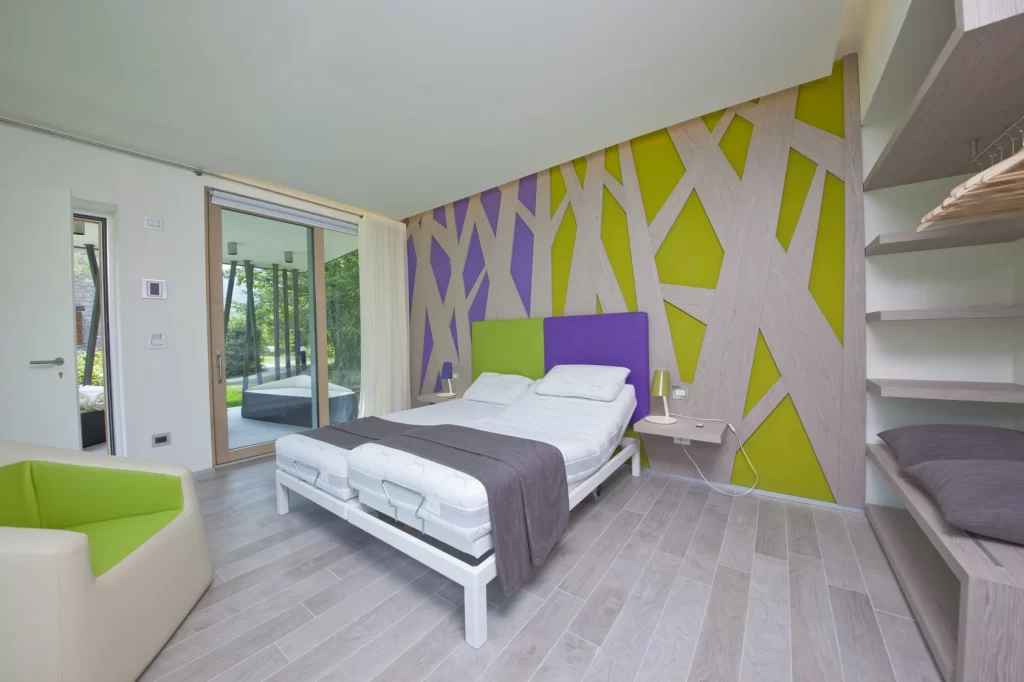 Camera d'hotel moderna con pareti decorate in legno chiaro, verde e viola, letto con cuscini bianchi, finestra panoramica, poltrona verde e comodini con lampade.