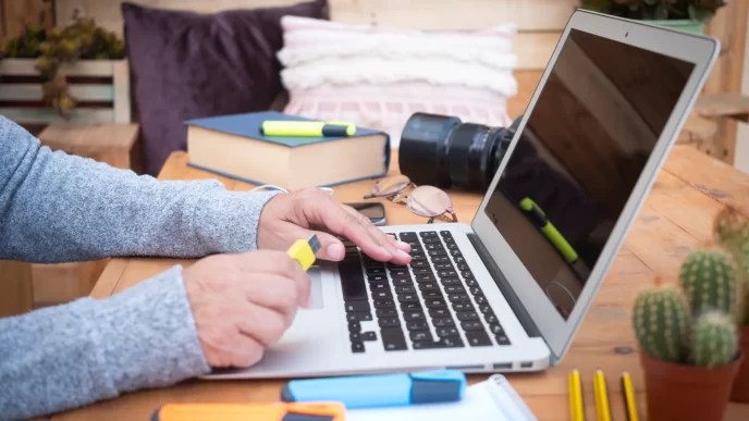 Donna che lavora su laptop a un tavolo di legno con libri, fotocamera e evidenziatori