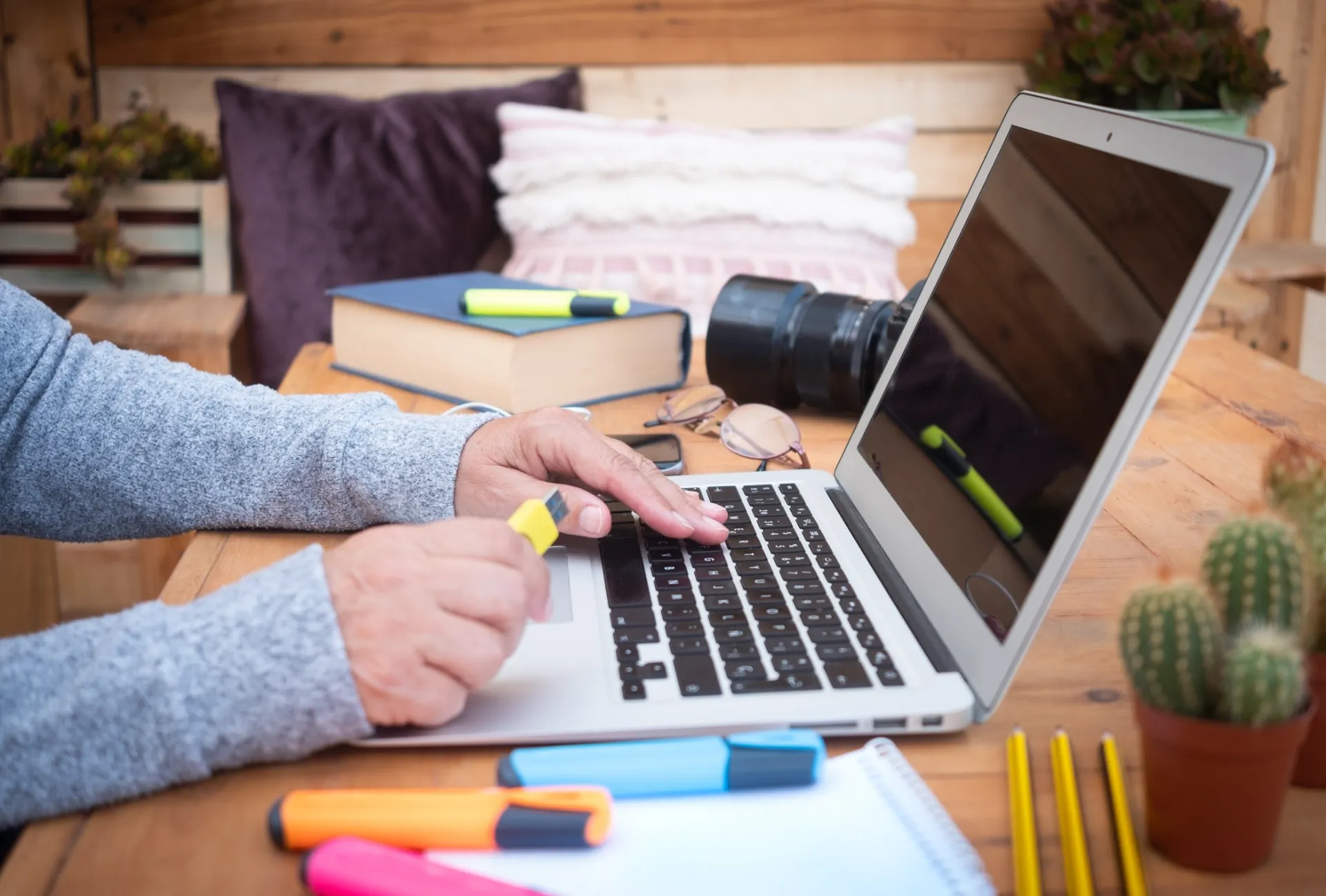 Donna che lavora su laptop a un tavolo di legno con libri, fotocamera e evidenziatori