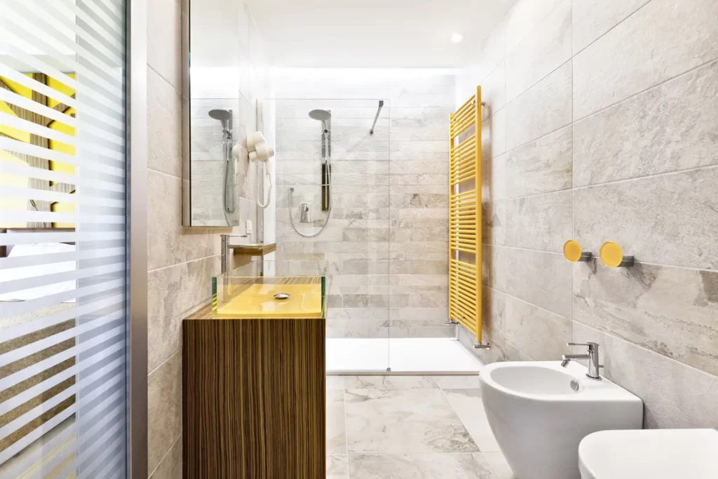 Bagno moderno di una camera d'hotel con doccia a filo pavimento, lavabo giallo, pareti in pietra grigia, asciugamani bianchi, bidet e radiatore giallo.