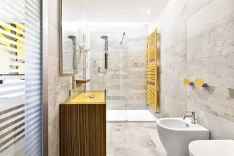 Bagno moderno di una camera d'hotel con doccia a filo pavimento, lavabo giallo, pareti in pietra grigia, asciugamani bianchi, bidet e radiatore giallo.