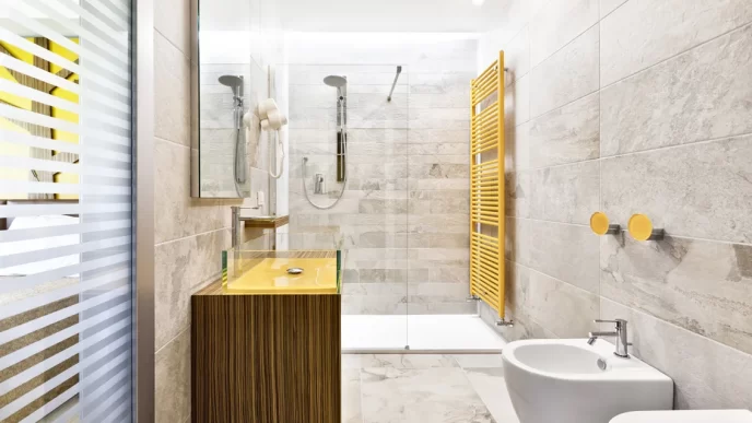 Bagno moderno di una camera d'hotel con doccia a filo pavimento, lavabo giallo, pareti in pietra grigia, asciugamani bianchi, bidet e radiatore giallo.