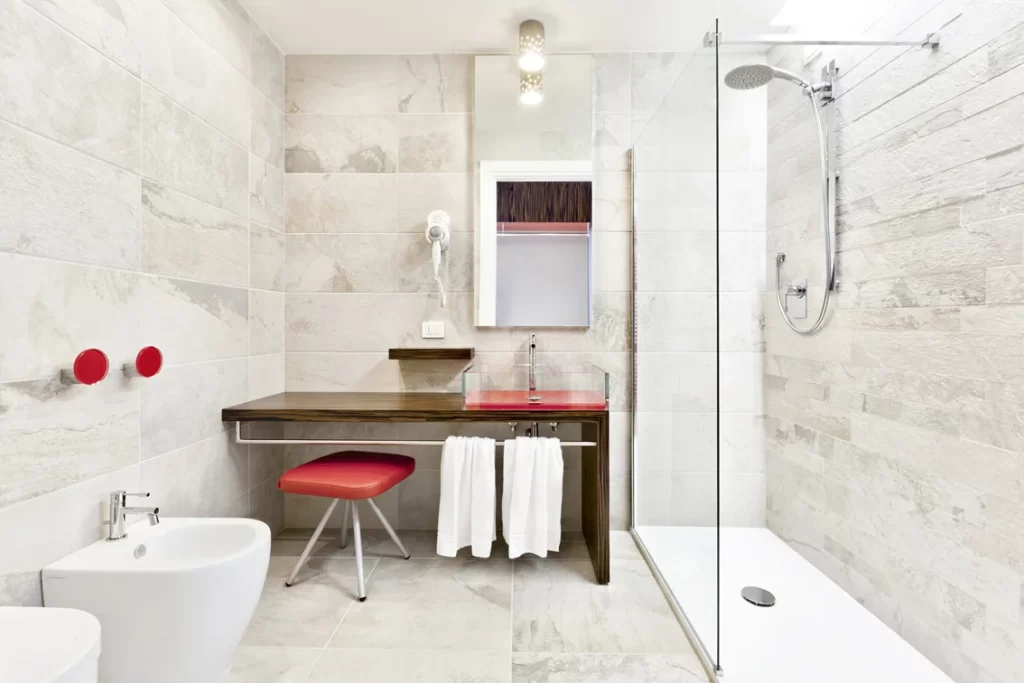 Bagno moderno di una camera d'hotel con doccia a filo pavimento, lavabo rosso, pareti in pietra grigia, bidet, sgabello rosso e asciugamani bianchi.