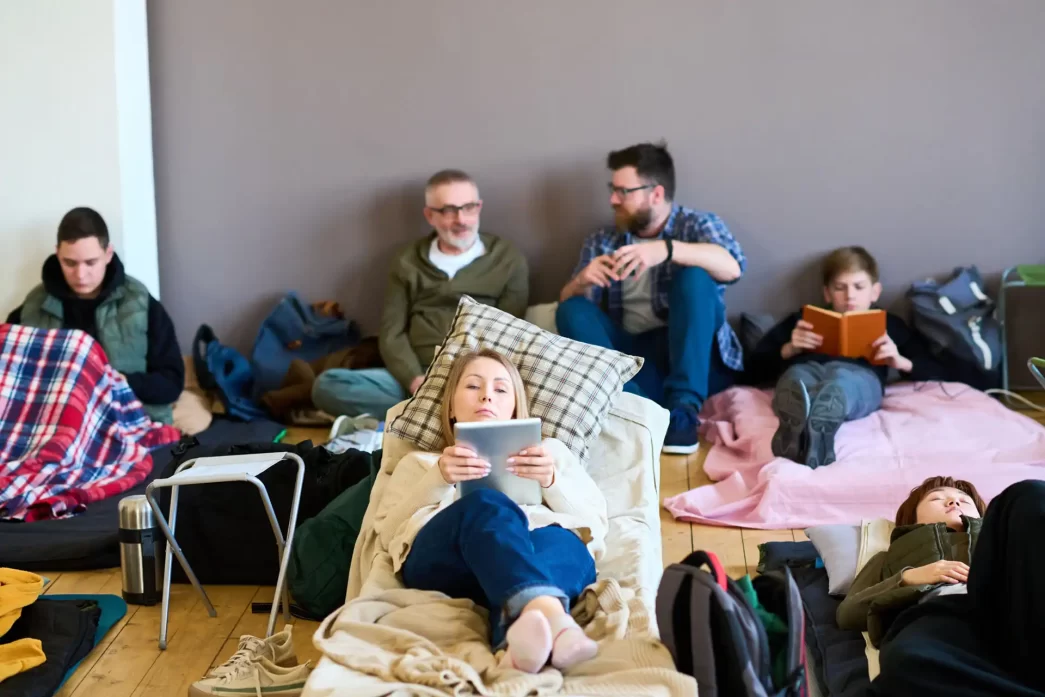 Gruppo di persone riposando in una stanza con letti improvvisati e coperte, una donna al centro usa un tablet.
