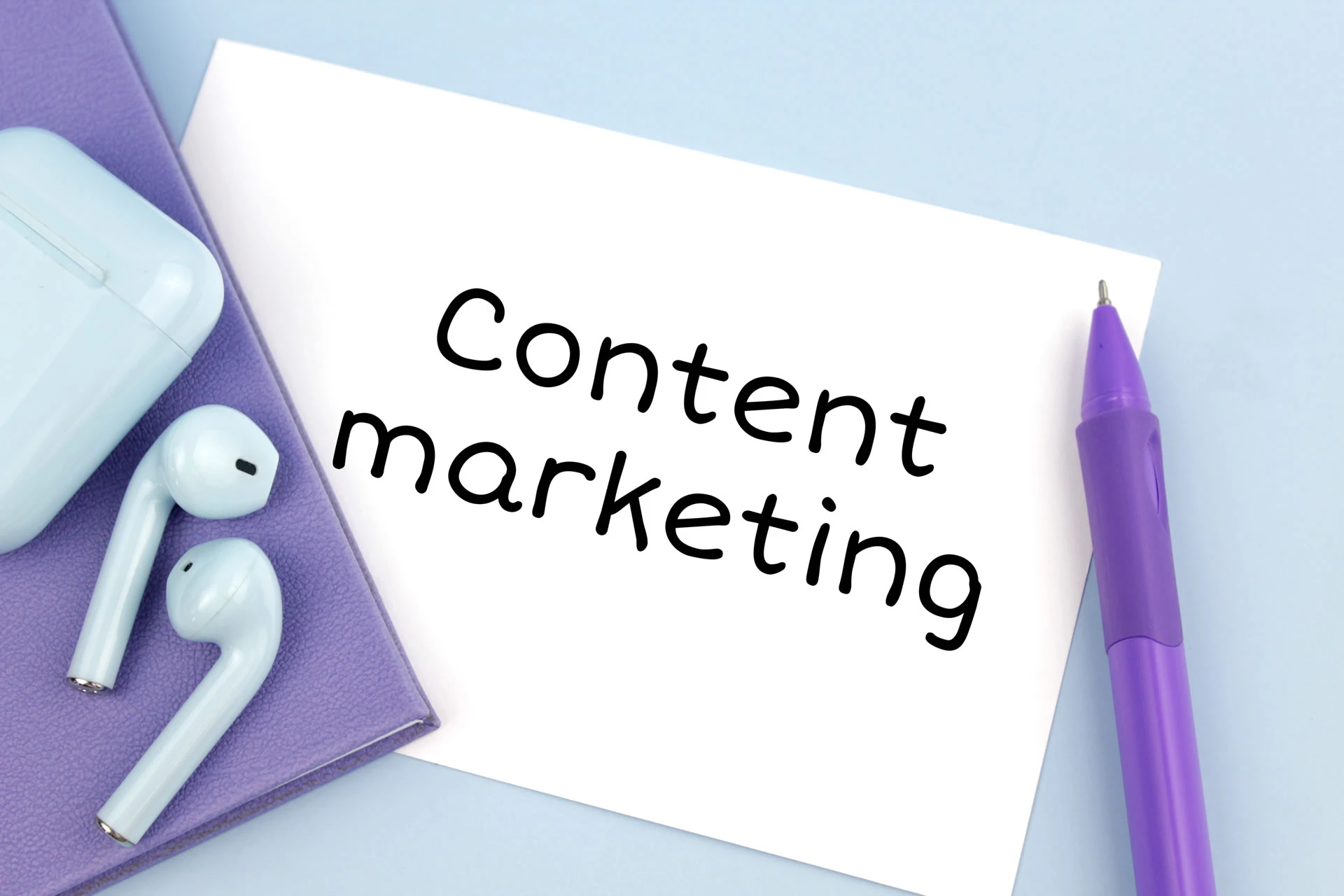 AirPods bianchi e penna viola su quaderno viola con nota 'Content marketing'