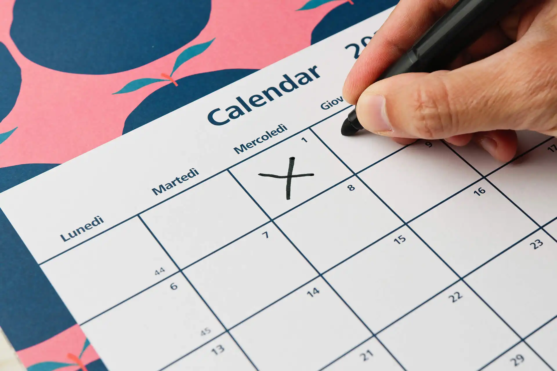 Primo piano di una mano che segna una data sul calendario, indicando il primo giorno del mese con una X.
