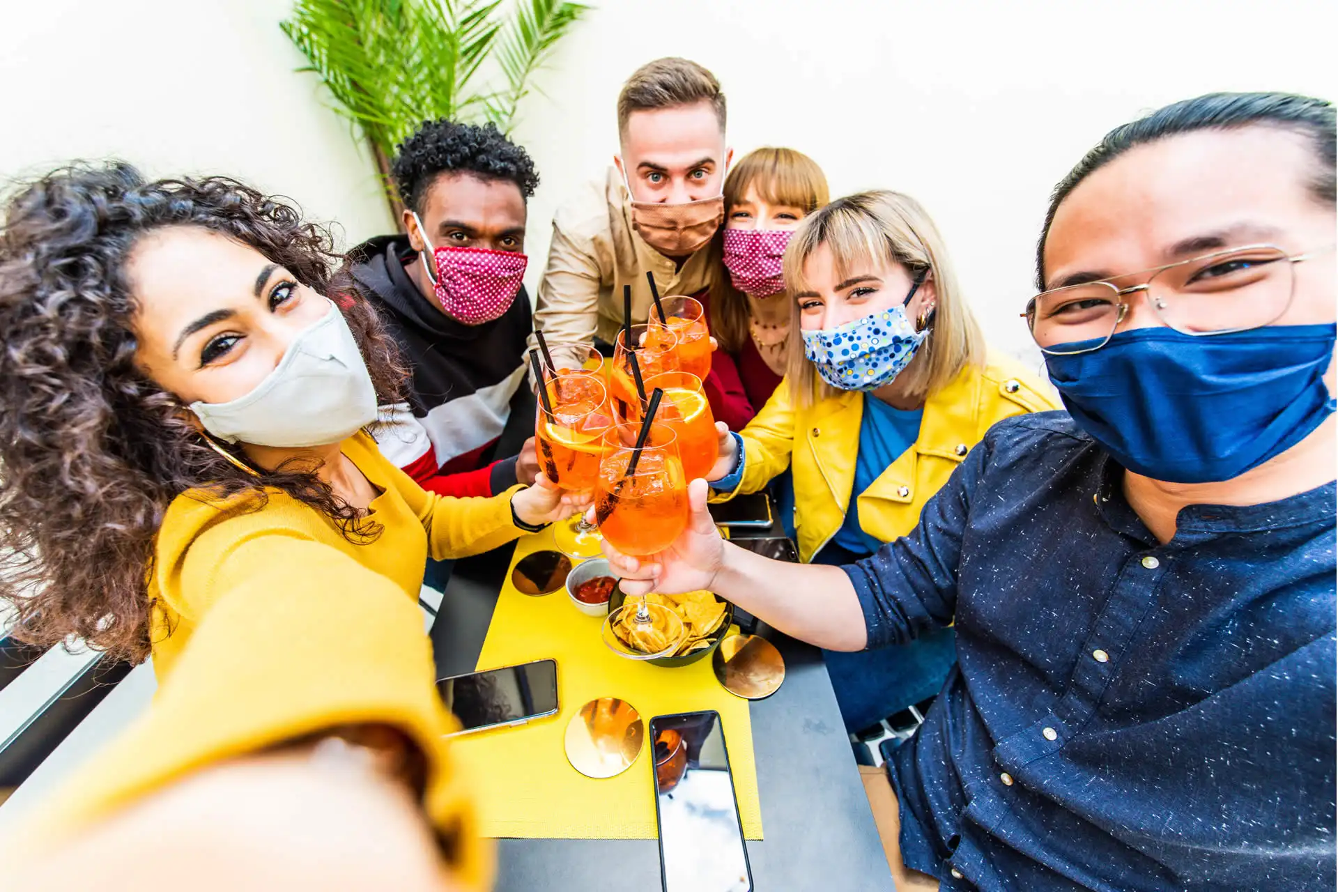 Un gruppo di amici multirazziali indossa mascherine protettive e brinda con cocktail durante l'aperitivo.