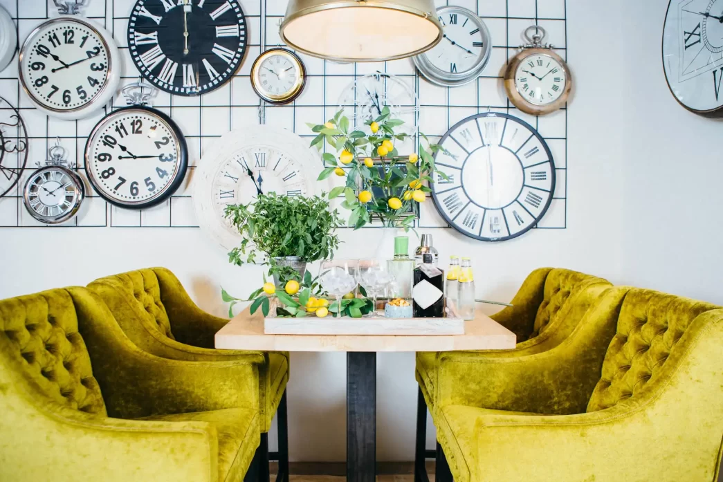 Interno di una zona pranzo elegante con sedie verde oliva e una parete decorata con vari orologi vintage.