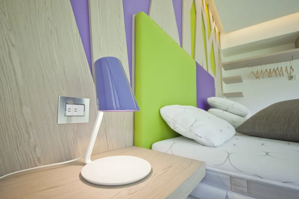Camera d'hotel moderna con decorazioni verde e viola, lampada da tavolo viola, letto con cuscini bianchi e pareti in legno chiaro.