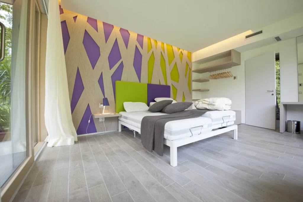 Camera d'hotel moderna con pareti decorate in legno chiaro e colori vivaci, letto con cuscini bianchi e grigi, lampada da tavolo viola e ampie finestre.