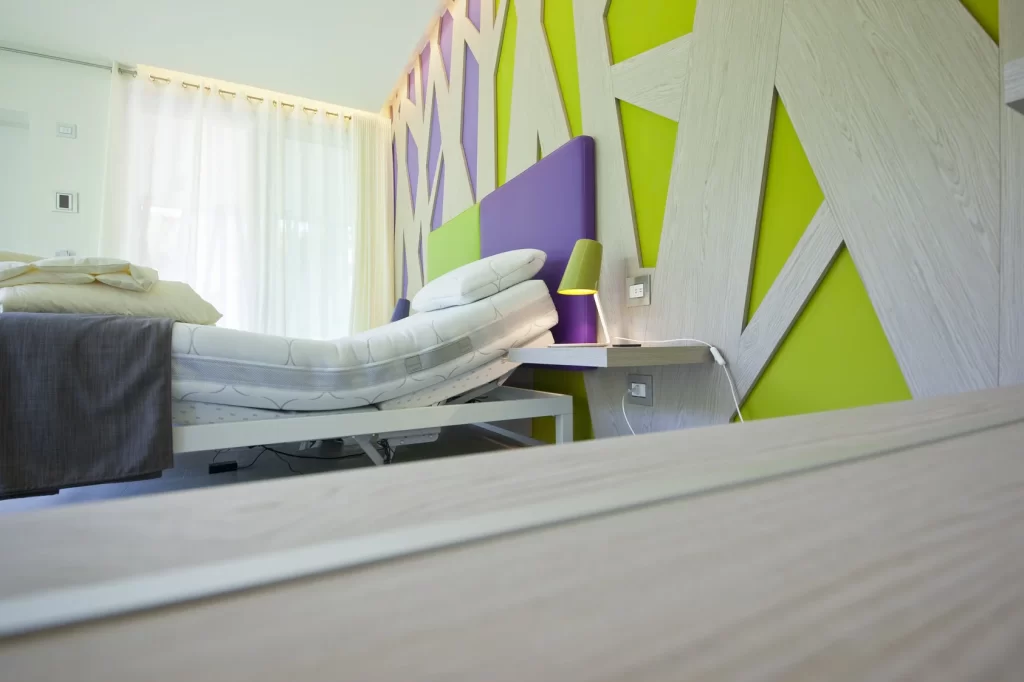 Camera d'hotel moderna con letto regolabile, pareti decorate in legno chiaro, verde e viola, tenda bianca, lampada da tavolo verde e arredamento minimalista.