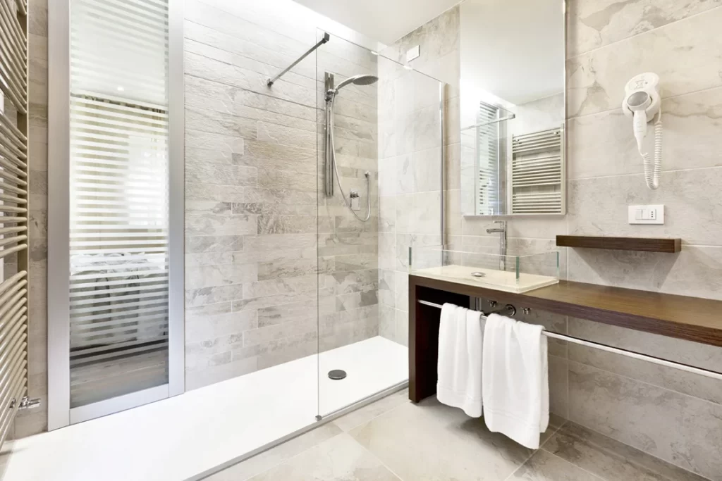 Bagno moderno di una camera d'hotel con doccia a filo pavimento, pareti in pietra grigia, lavabo con specchio, phon e asciugamani bianchi.
