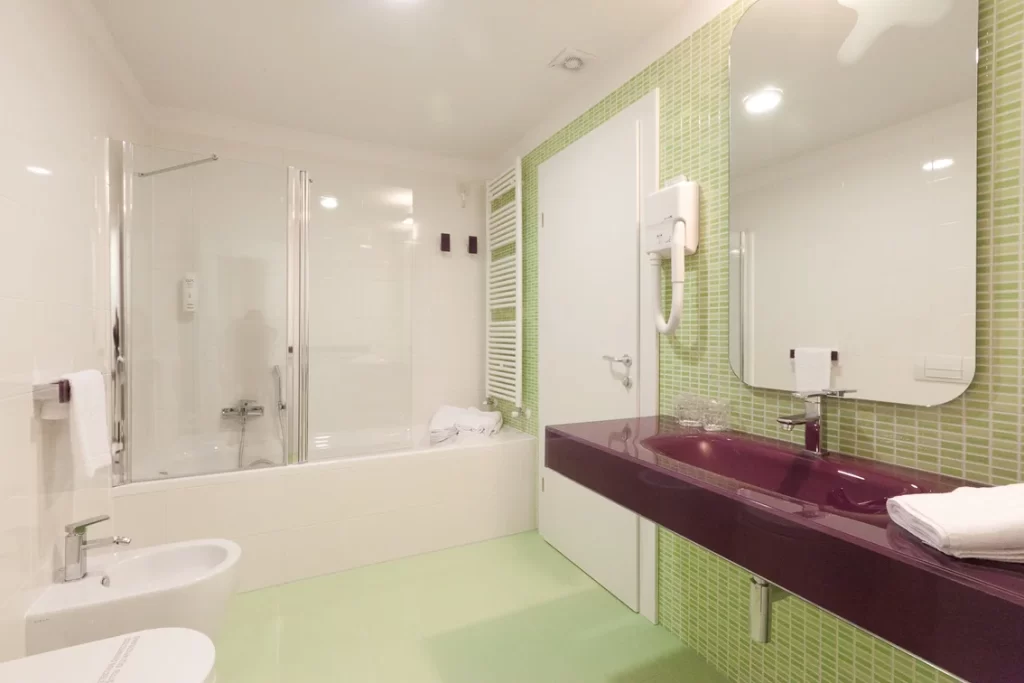 Bagno moderno di una camera d'hotel con vasca, pareti bianche, lavabo viola, piastrelle verdi, bidet, phon a muro e asciugamani bianchi.