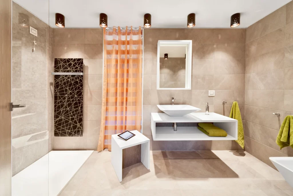 Bagno moderno di una camera d'hotel con doccia a filo pavimento, lavabo sospeso bianco, pareti in piastrelle beige, asciugamani verdi e tenda arancione.
