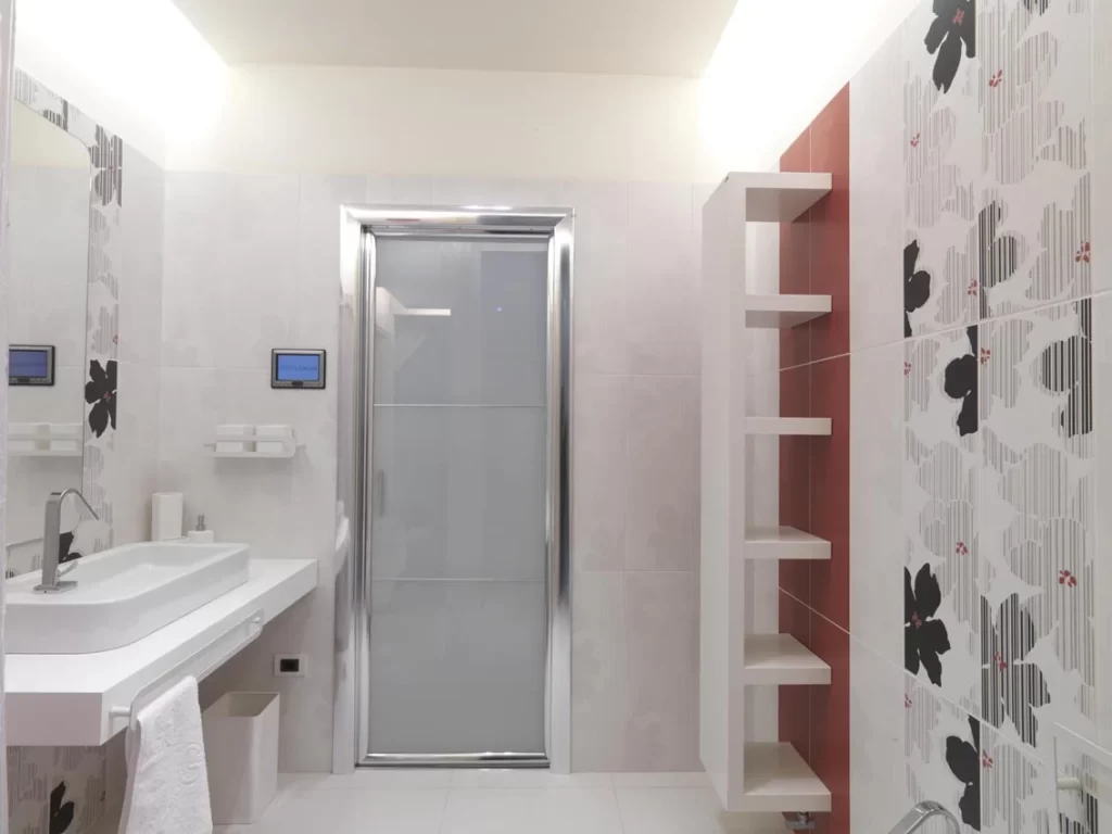 Bagno moderno di una camera d'hotel con doccia, lavabo bianco, specchio con decori floreali, piastrelle bianche e nere, mensole aperte e asciugamani bianchi.