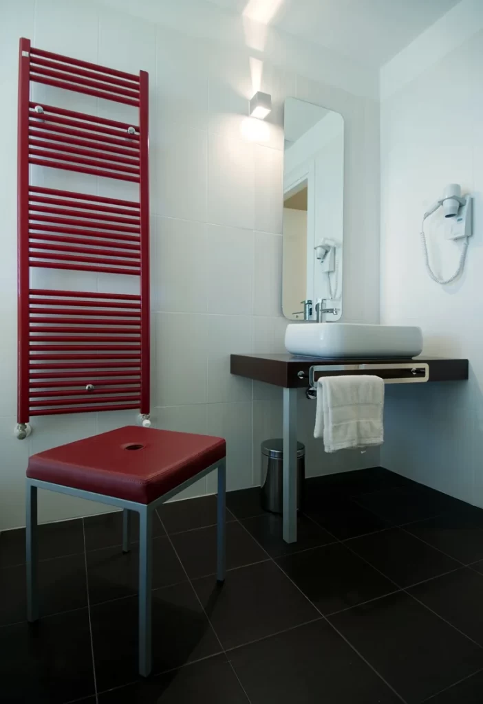 Bagno moderno di una camera d'hotel con arredi minimalisti, lavabo bianco, pareti bianche, radiatore rosso, sgabello rosso e phon a muro.