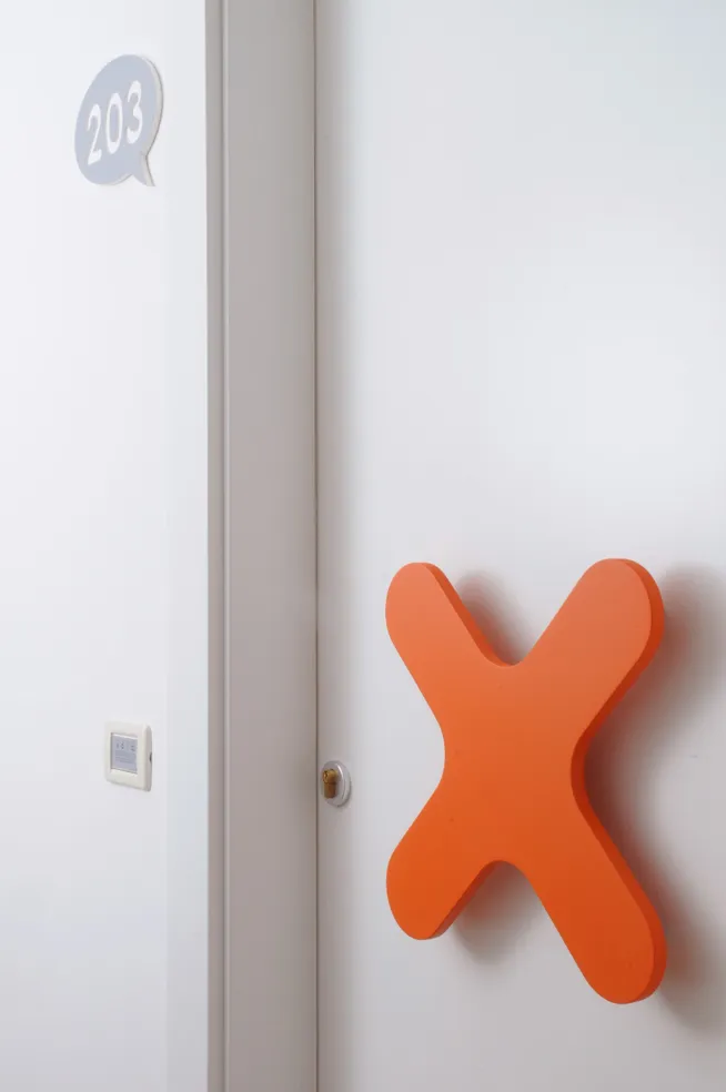 Primo piano della porta della camera d'hotel con grande maniglia a forma di X arancione.