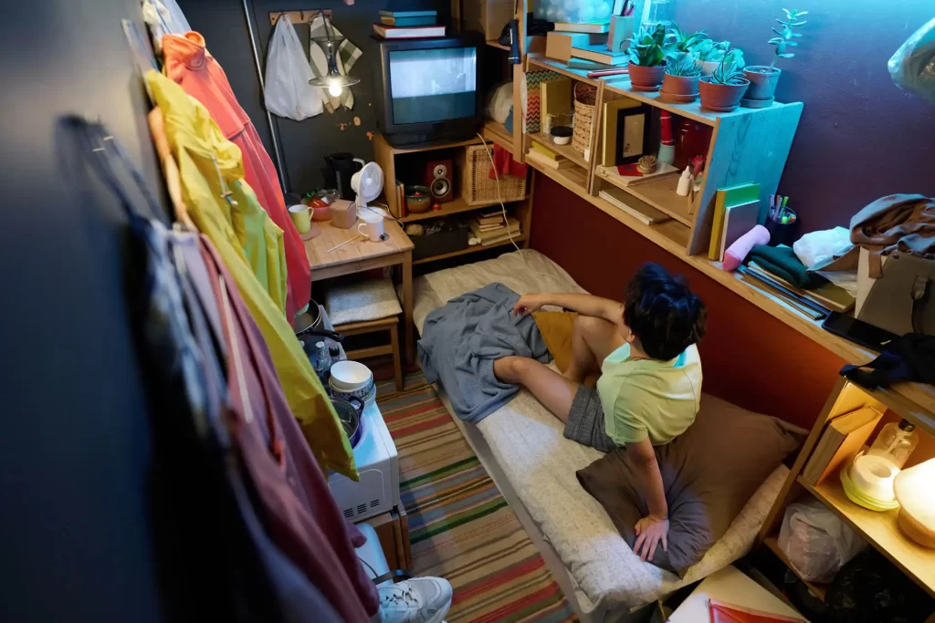 Persona seduta su un letto in una piccola camera senza finestre, arredata con scaffali, piante e una TV.