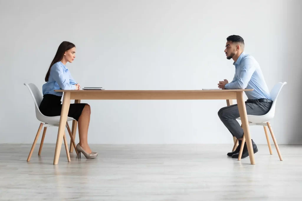 Due persone sedute distanti a un tavolo durante un incontro formale in una stanza minimalista.
