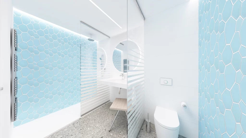 Bagno moderno di un hotel con piastrelle esagonali azzurre, specchio rotondo, lavabo bianco, doccia a vetri e pavimento in granito chiaro.